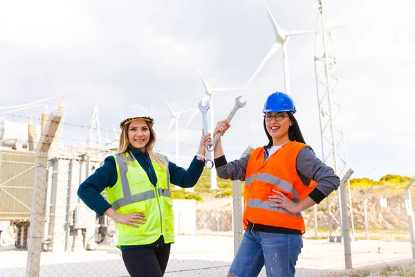 Portrait of happy two young technical engineers woman looking at camera posing and smiling while flexing her biceps with metal english key against of electrical components and wind turbines of an electricity power generation station