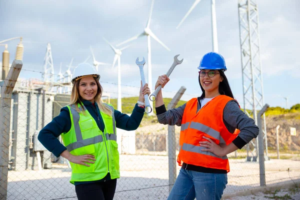 Portrait of happy two young technical engineers woman looking at camera posing and smiling while flexing her biceps with metal english key against of electrical components and wind turbines of an electricity power generation station