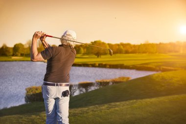 Kıdemli golf oyuncusu gün batımında, arka planda göl ile top çekim.