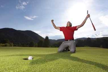 Profesyonel genç erkek golf oyuncusu diz ve kollar golf yeşil elinde putter ile kaldırdı golf topu fincan içine düşer gibi overjoyed olmak.