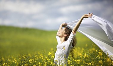 Sarı kolza tarlasında duran genç mutlu kadın rüzgarda beyaz bir bez parçası yla özgürlüğünü ifade ediyor.