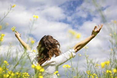 Young woman standing in yellow rapeseed field raising her arms expressing gratitude or freedom, view from behind. clipart