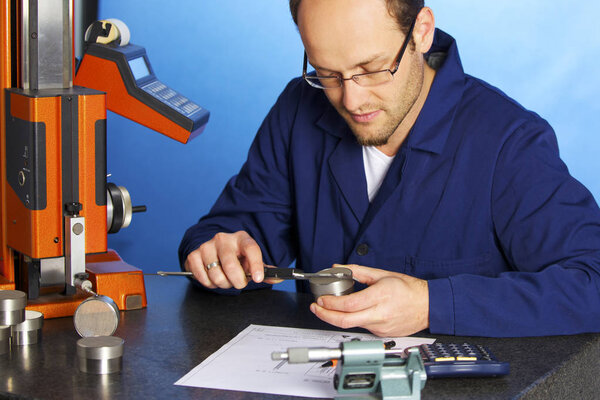 Young male engineer in blue overall measuring a metal part with caliper, isolated on blue background.