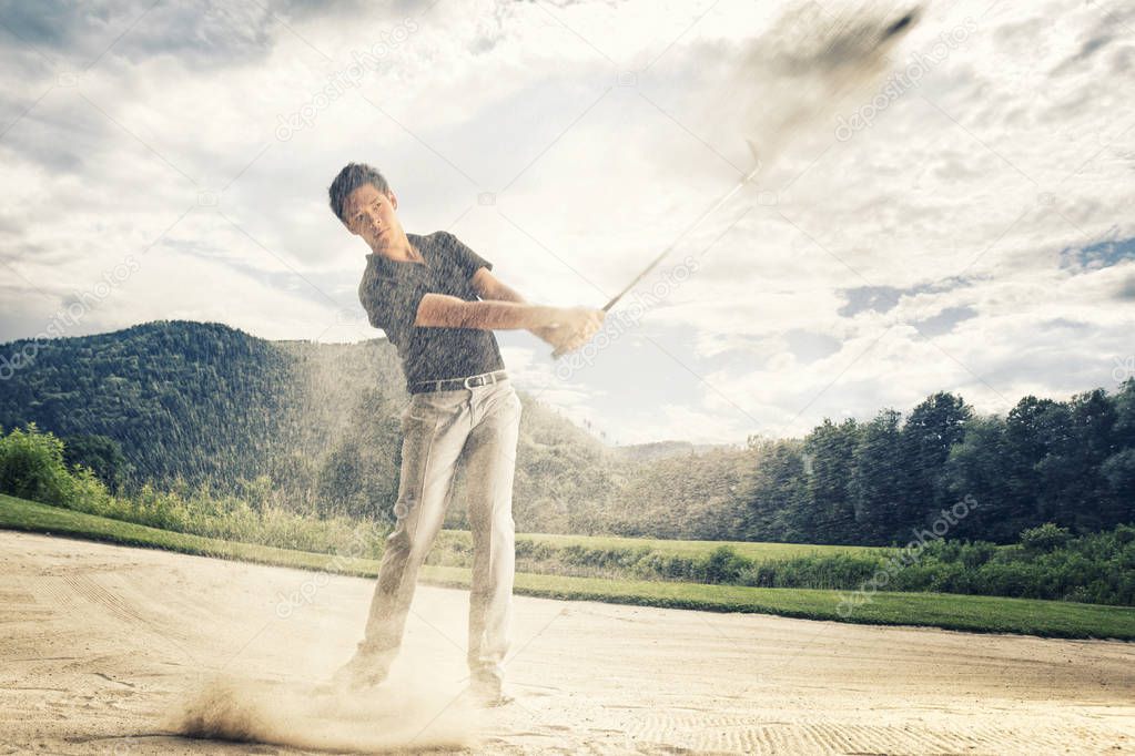 Male golf player in blue shirt and grey pants hitting golf ball out of a sand trap with sand wedge and sand caught in motion.