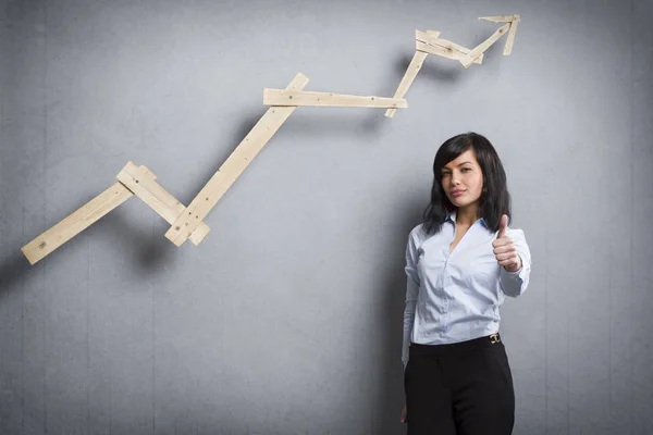 Concept: Successful career or business. Young confident businesswoman holding thumb up in front of ascending business graph, isolated on grey background.