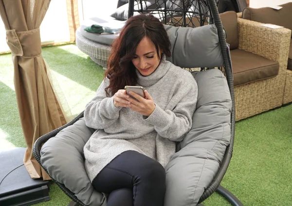 Young woman sitting in the cosy egg chair and looking into the smart phone and smiling