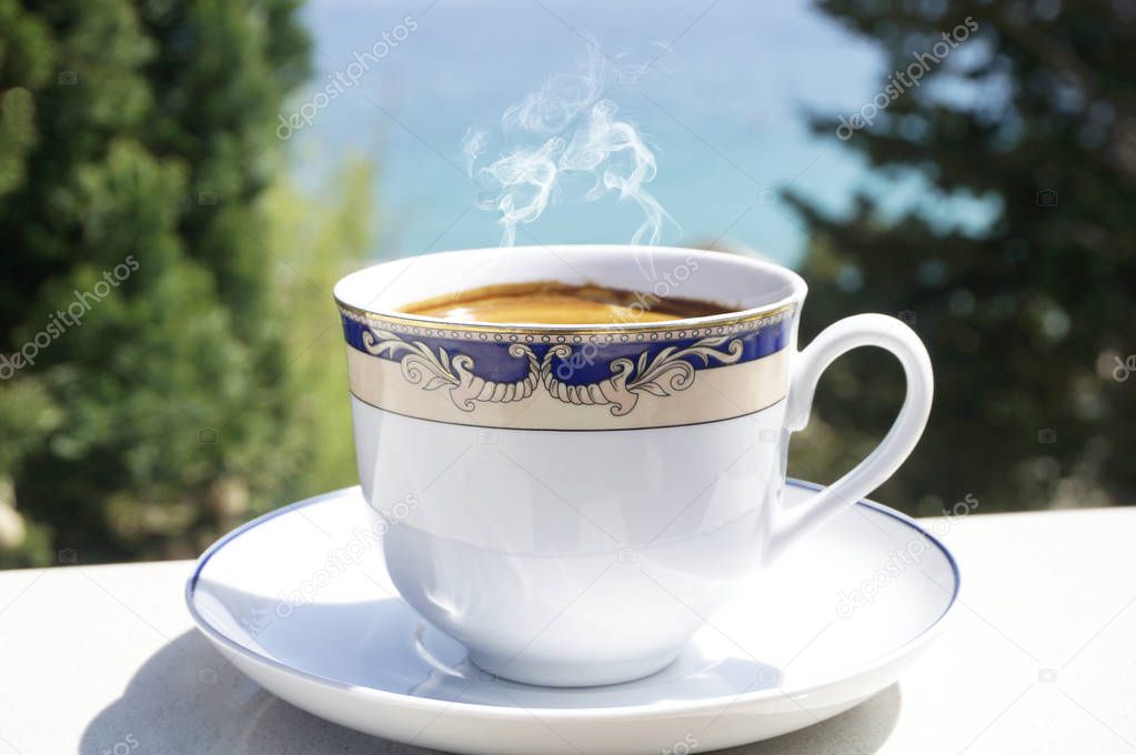 Cup of hot coffee on terrace with blurred sea and trees in the background