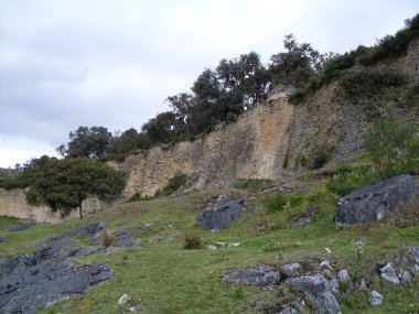 Kuelap kalesinin ana girişinin solundaki duvar, katı taş, bitki örtüsü ve bulutlu gökyüzü üzerine inşa edilmiş.