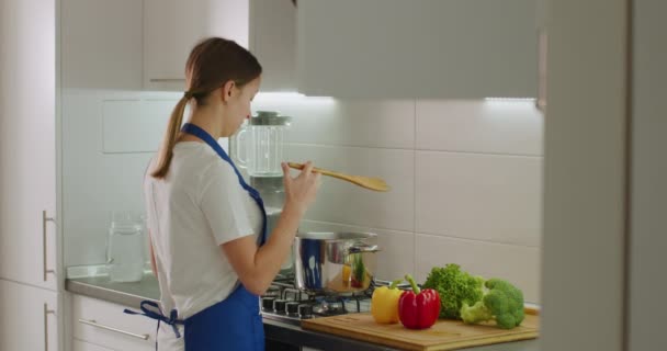 La femme prend une spatule en bois et mélange la vaisselle bouillante à la casserole sur le poêle. 4k — Video