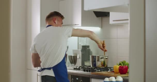 A man is listening to music and mixing boiling dishes at pan on the stove. 4k — Stock Video