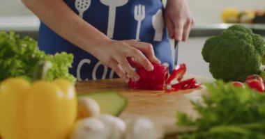 Yakın plan çekim. Bir kadın mutfak tahtasında büyük bir biber dilimliyor. Etrafta bir sürü sebze var. 4K