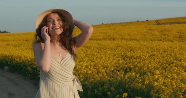 La ragazza risponde al telefono con sorpresa e parla felicemente. Una ragazza sorridente sta attraversando il campo giallo. Tramonto. 4k — Video Stock