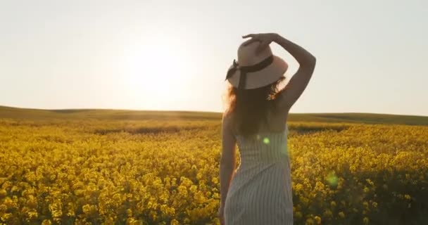 Uma linda garota está andando ao longo do campo e colocando seu chapéu. Campo com flores amarelas. Pôr-do-sol ao fundo. 4k — Vídeo de Stock
