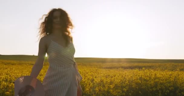 A menina sorrindo está tirando o chapéu e girando ao redor. Campo com flores amarelas. Pôr-do-sol ao fundo. 4k — Vídeo de Stock