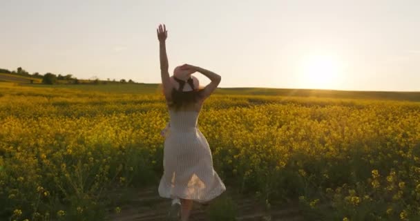 Gülümseyen bir kız sahada koşuyor ve şapkasını elinde tutuyor. Durup gün batımını seyrediyor. Sarı çiçekli tarla. 4k — Stok video