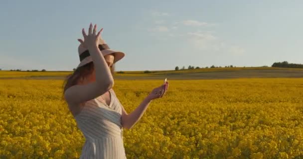 Uma rapariga sorridente está a atravessar um campo amarelo. A rapariga está a ajustar o chapéu e a olhar-se ao espelho. Pôr do sol. 4k — Vídeo de Stock