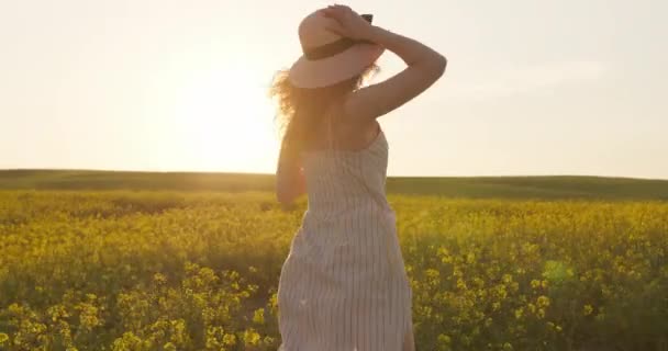 Uma menina sorrindo está correndo pelo campo e segurando seu chapéu. Campo com flores amarelas. Pôr-do-sol ao fundo. 4k — Vídeo de Stock