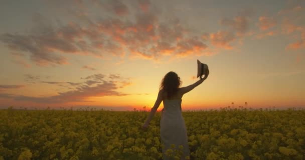 Девушка стоит среди поля желтых цветов и надевает шляпу. Девушка смотрит на закат. 4K — стоковое видео