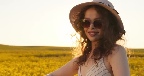Záběr zblízka. Usmívající se dívka ve slunečních brýlích se dívá do kamery. Slunce svítí v pozadí a žluté květy. 4K — Stock video