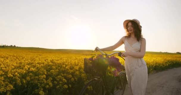 Dívka kráčí po polní cestě s bicyklem. Krásné retro kolo s košíkem květin. Západ slunce v pozadí. 4K — Stock video