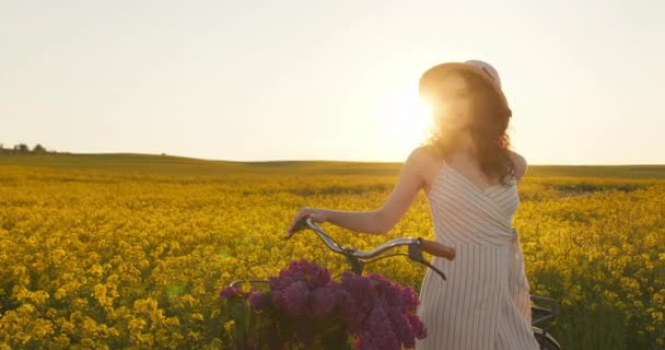 Dziewczyna stoi i opiera się o rower. Patrzy w kamerę i uśmiecha się. Słońce świeci w tle. Podnosi rękę i trzyma kapelusz. 4K — Wideo stockowe