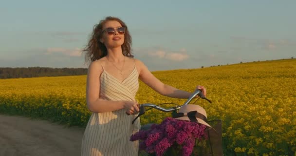 Ένα κορίτσι περπατάει σ 'ένα δρόμο με ποδήλατο. Φοράει φόρεμα και γυαλιά ηλίου. Όμορφο ρετρό ποδήλατο με καλάθι με λουλούδια. 4K — Αρχείο Βίντεο