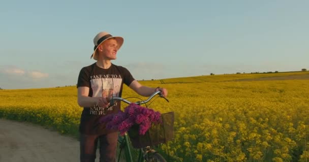 Chlápek kráčí po polní cestě s bicyklem. Má na sobě dámskou čepici. Krásné retro kolo s košíkem květin. 4K — Stock video