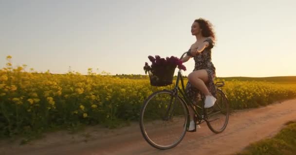 Dívka na kole jede po polní cestě poblíž pole žlutých květin. Retro kolo s košíkem květin. 4K — Stock video