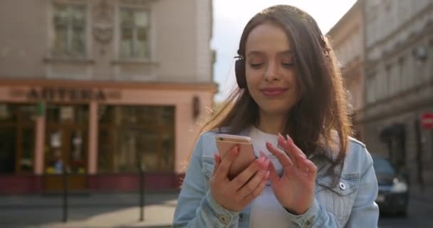 Disparos de cerca. La chica está enviando mensajes de texto y escuchando música en auriculares grandes. El pelo vuela en el viento. 4K — Vídeos de Stock