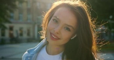 Yakın plan çekim. Kameraya bakan bir kızın yüzü gibi. O gülümsüyor ve küçük kulaklıklarla müzik dinliyor. Saçları rüzgarda dalgalanıyor. 4K