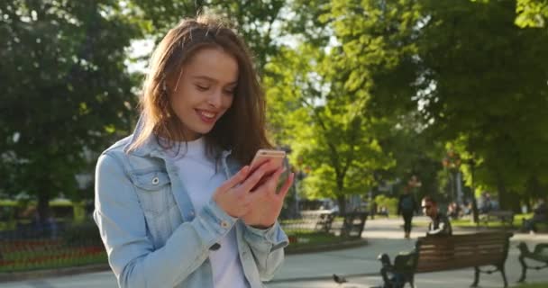 Záběr zblízka. Veselá holka stojí a píše do telefonu. Směje se a poslouchá hudbu s malými sluchátky. Vlasy se jí třepotají ve větru. Letní slunce svítí. 4K — Stock video