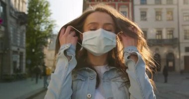 Kız koruyucu maskesini çıkarıyor ve gülümsüyor. Özgür olmanın sevinci. Saçları rüzgarda dalgalanıyor. Yaz güneşi parlıyor. Arka planda şehrin merkezi. 4K