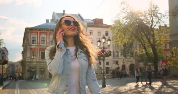 Una chica sonriente está caminando y hablando emocionalmente por teléfono. Caminando con un clima soleado por la ciudad. 4K — Vídeos de Stock