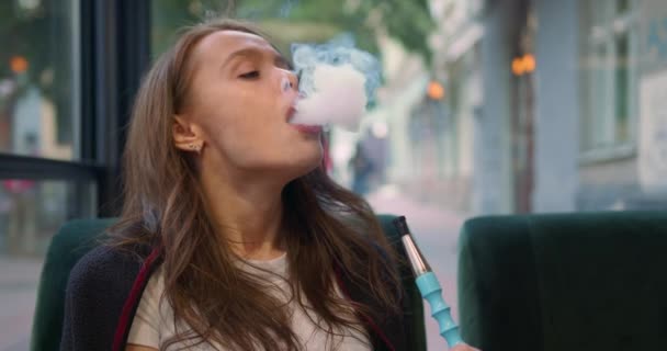 Nahaufnahme. Das Mädchen sitzt in einem Café auf der Veranda. Sie raucht eine Wasserpfeife und gibt langsam Rauch ab. Dann blickt sie in die Kamera und lacht. 4K — Stockvideo