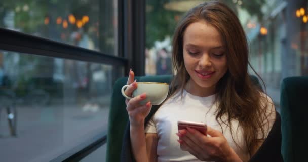 Disparos de cerca. La chica está sentada en un café en la terraza. Está bebiendo té y mandando mensajes por teléfono. La chica sonriente está envuelta en una manta. 4K — Vídeos de Stock