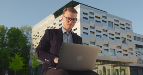 Een zakenman rondt zijn werk op een laptop af. Een man zit in een park in de buurt van een zakencentrum. Een lachende man met een bril. 4K — Stockvideo