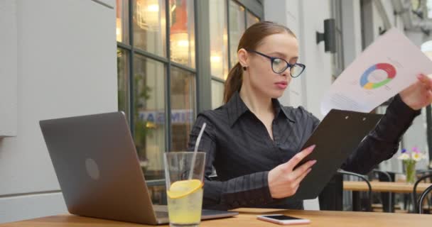 La fille regarde à travers les documents de travail avec des diagrammes. Une femme d'affaires travaille sur la véranda d'un café. Il y a de la limonade sur la table et un ordinateur portable. Elle portait des lunettes et une chemise. 4K — Video