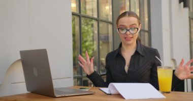 Bir iş kadını video görüşmelerinde iş konularını tartışıyor. Duygusal olarak açıklama yapıyor ve ellerini sallıyor. O, bir kafede bir masada oturuyor. Masada limonata ve dizüstü bilgisayar var. 4K