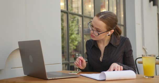 Bizneswoman pracuje nad laptopem. Starannie wprowadza dane z dokumentów. Siedzi przy stole w kawiarni. 4K — Wideo stockowe