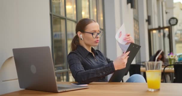 İş kadını çalışan belgelere dikkatlice bakıyor. O, bir kafede bir masada oturuyor. Masada limonata ve dizüstü bilgisayar var. 4K — Stok video