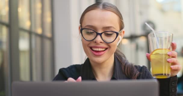 Yakın plan çekim. Mutlu bir iş kadını gülümsüyor ve bir dizüstü bilgisayar üzerinde çalışıyor. Limonatasını içiyor. O, bir kafede bir masada oturuyor. 4K — Stok video