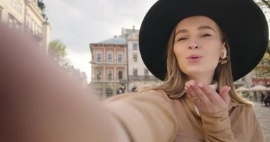 Yakın plan çekim. Gülümseyen bir kız ön kamerada bir video çekiyor. Kız kameraya bir öpücük veriyor. Güneş parlıyor ve insanlar arka planda yürüyorlar. 4K