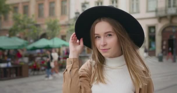 Disparos de cerca. La chica está mirando a la cámara y sonriendo. Ella sostiene su sombrero en su mano. Las gotas de lluvia pasan volando. La plaza central de la ciudad en el fondo. 4K — Vídeos de Stock