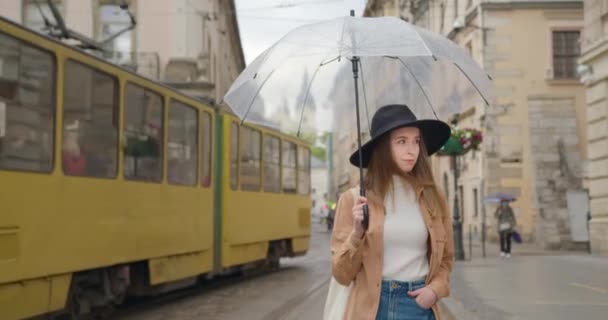 少女はゆっくりと雨の中を歩いている。彼女は傘を手に持っている。少し雨が降っている。彼女は頭に大きな帽子をかぶっている。後ろに路面電車が通っている。4K — ストック動画