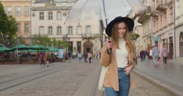 女の子は傘を開けて笑っている。彼女は回転している。少し雨が降っている。彼女は頭に大きな帽子をかぶっている。彼女は街の中央広場を歩いている。4K — ストック動画