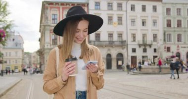 Kız telefonda mesajlaşıyordu. Yürüyor ve gülümsüyor. Elinde bir fincan kahve tutuyor. Büyük bir şapka takıyor. Güneş parlıyor ve insanlar arka planda yürüyorlar. 4K