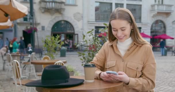 A menina está sentada em uma mesa em um café e mensagens de texto no telefone. Na mesa há uma chávena de café e um chapéu. A praça central da cidade ao fundo. 4K — Vídeo de Stock