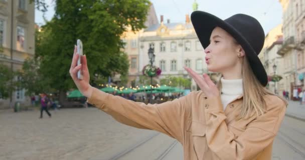 Una chica sonriente está tomando un video en la cámara frontal. La chica está haciendo un beso a la cámara. Lleva un gran sombrero negro. El sol brilla y la gente camina en el fondo. 4K — Vídeos de Stock