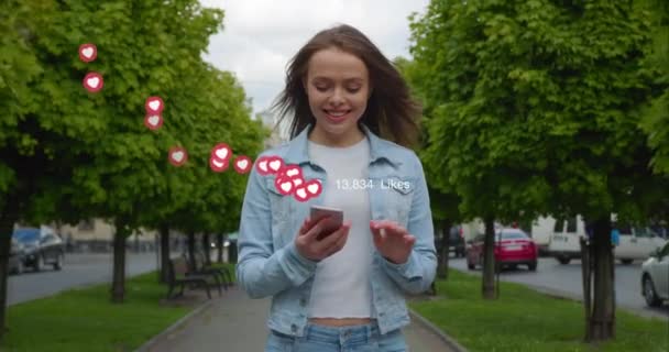 La chica está caminando por el callejón y mirando sus redes sociales. Ella está contenta con el número de me gusta que recibió. Su pelo está revoloteando en el viento. Árboles y personas en el fondo — Vídeos de Stock