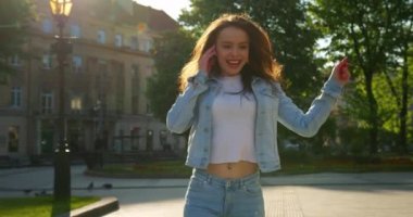 Neşeli bir kız şarkı söylüyor ve dans ediyor. O gülüyor ve küçük kulaklıklarla müzik dinliyor. Saçları rüzgarda dalgalanıyor. Yaz güneşi parlıyor. 4K
